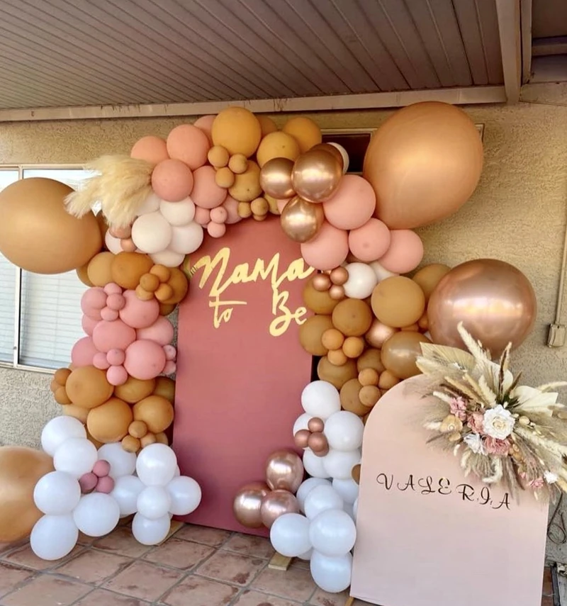 A baby-shower backdrop reading Mama to Be in blush and rose gold, framed by a balloon garland and pampas grass
