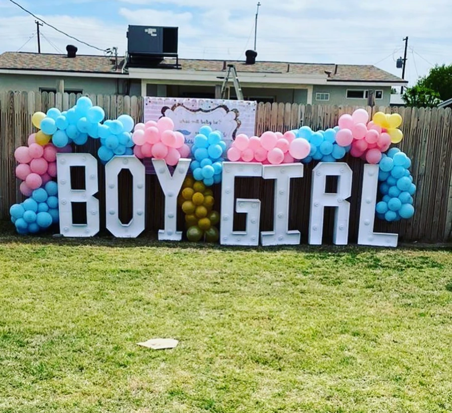 An outdoor gender-reveal display with freestanding BOY and GIRL letters and pink and blue balloon garlands