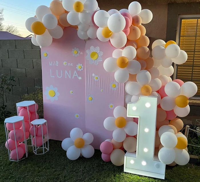 A first-birthday backdrop with a pastel arch, daisies, a lit number one and a balloon garland