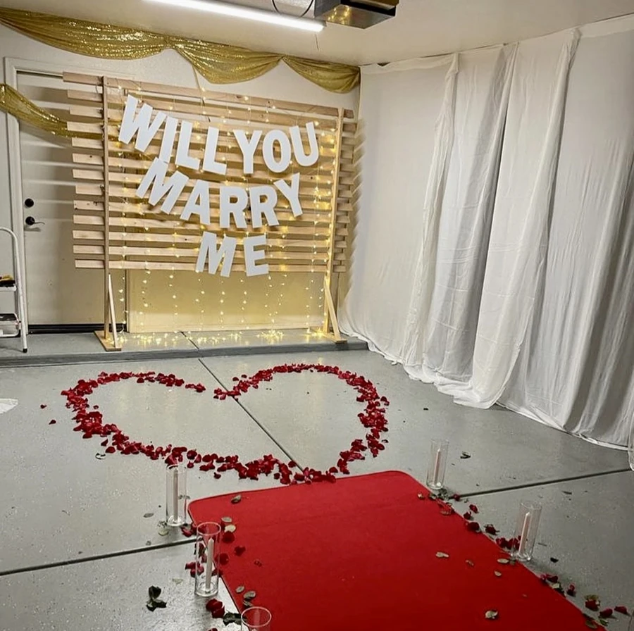 A proposal setup with a lit Will You Marry Me backdrop, a red carpet and a heart of rose petals
