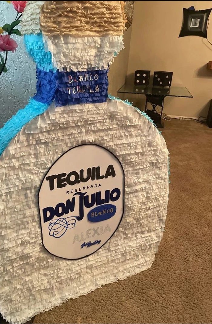 A custom pi&ntilde;ata shaped like a tall tequila bottle, taller than the chair beside it